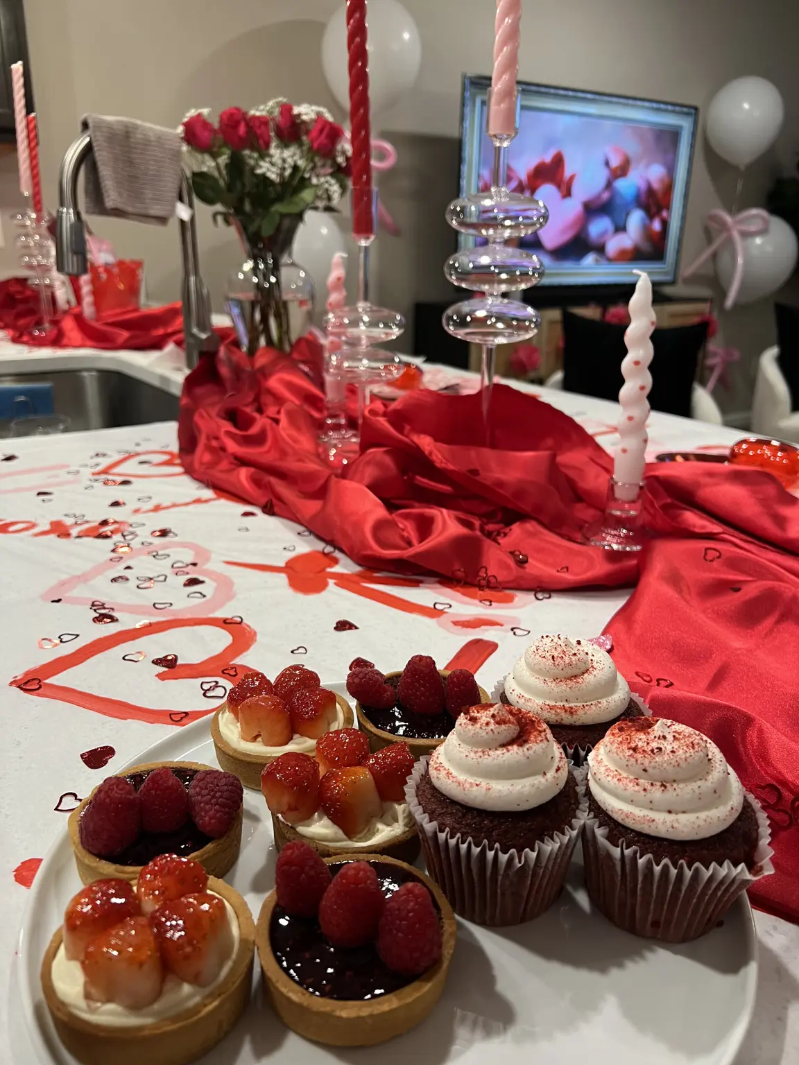 Berry tarts and red velvet cupcakes in front of a red satin runner and glass candleholders at a Galentine’s party